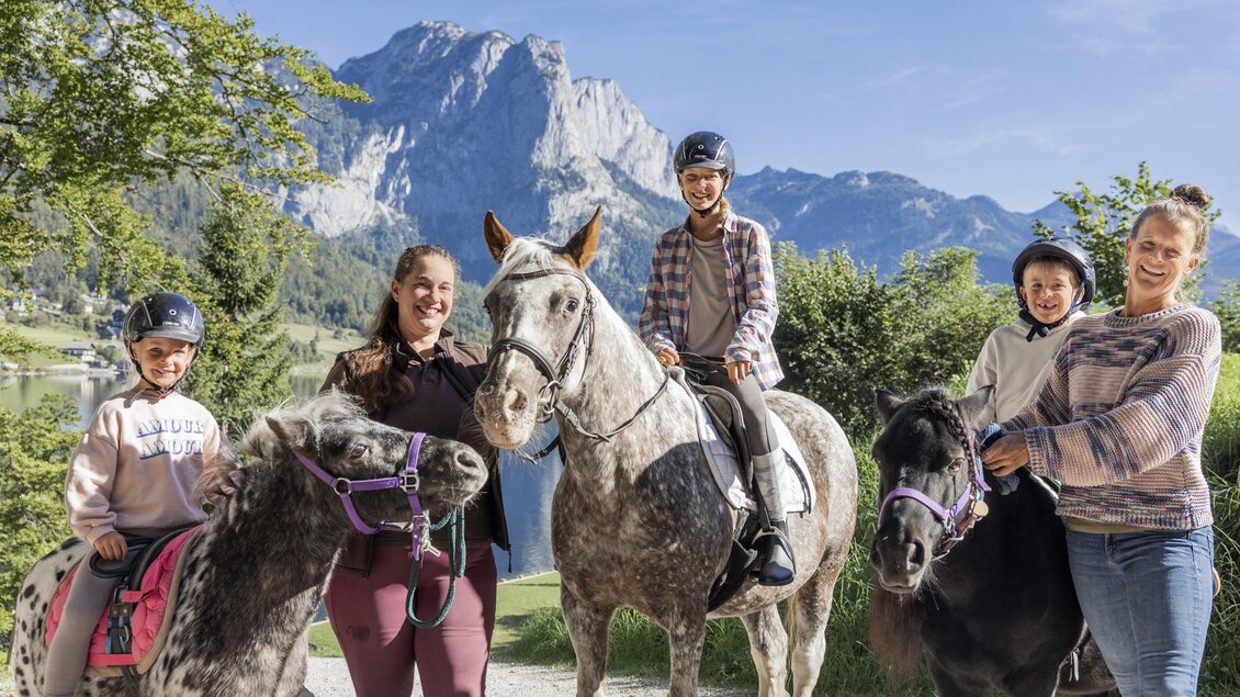 Eine Gruppe von Menschen steht mit Ponys und einem Pferd vor einer Berglandschaft. Die Kinder und Erwachsenen tragen Helme und lächeln in die Kamera. | © www.zloam.at