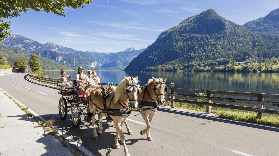 Eine Pferdekutsche fährt an einem malerischen See entlang, umgeben von Bergen. Die Sonne scheint und der Himmel ist klar. | © www.zloam.at