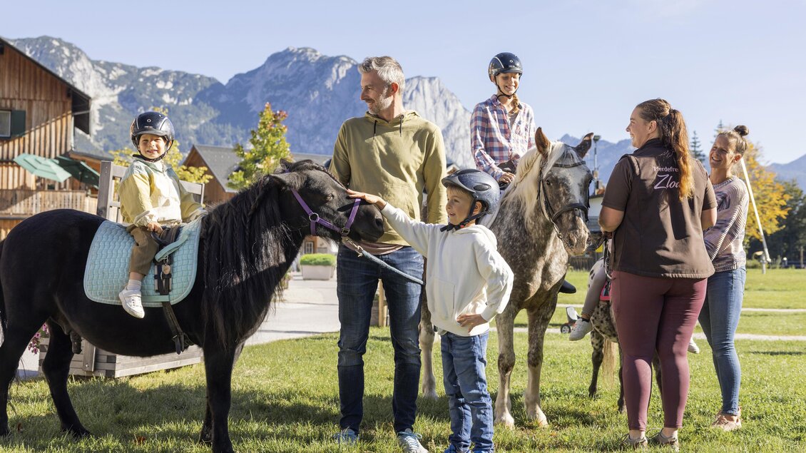 Eine Gruppe von Kindern und Erwachsenen steht im Freien in der Nähe von Ponys und Pferden. Im Hintergrund sind Berge und ein klarer Himmel zu sehen. | © www.zloam.at