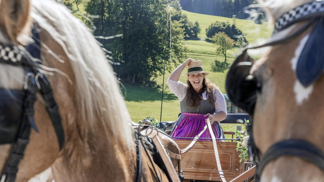 Eine Frau in traditioneller Kleidung fährt mit einer Pferdekutsche durch eine grüne Landschaft. Im Hintergrund sind Bäume und Hügel zu sehen. | © www.zloam.at