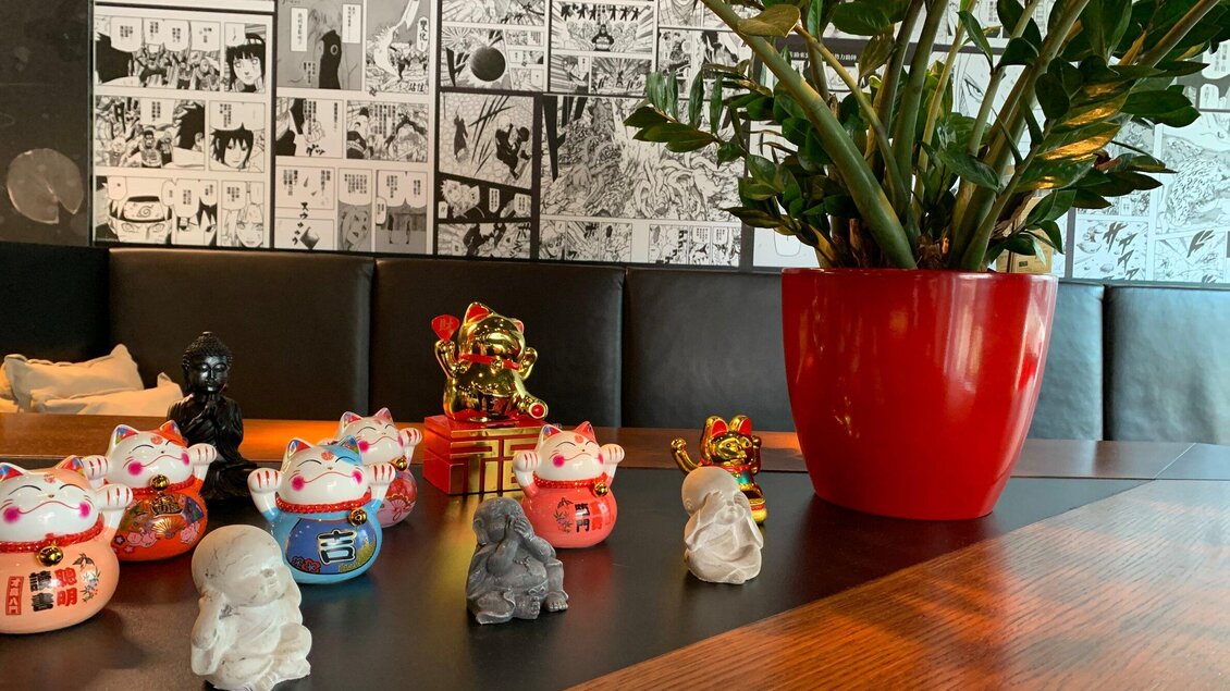 Verschiedene Glücksbringer-Figuren stehen auf einem Tisch, umgeben von einem großen roten Blumentopf. Im Hintergrund sind Manga-Illustrationen zu sehen. | © Pho You