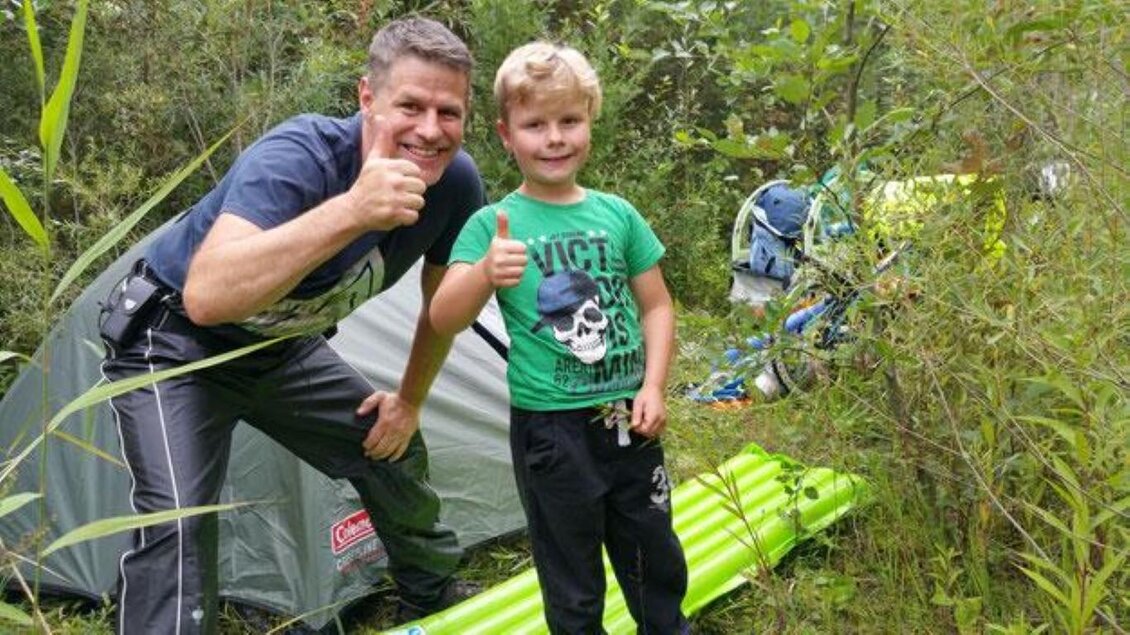 Ein Vater und sein Sohn stehen vor einem Zelt im Freien. Beide lächeln und zeigen einen Daumen nach oben. | © www.physiotherapie-neumeister.at