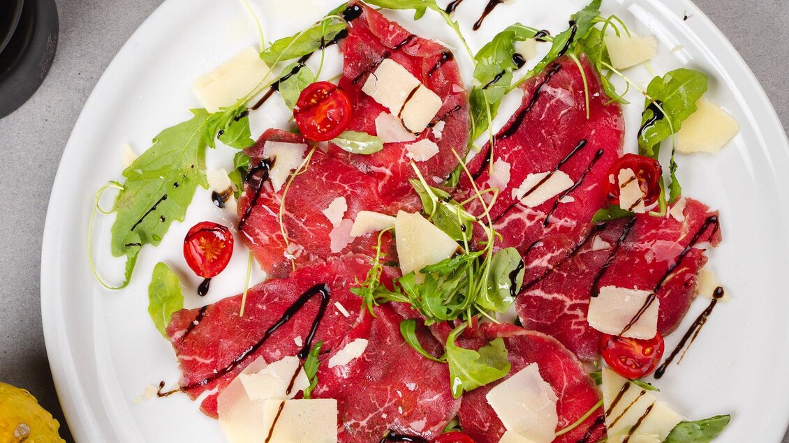 Ein Teller mit dünn geschnittenem Rindfleisch, garniert mit Rucola, Kirschtomaten und Parmesan. Darüber ist eine Balsamico-Reduktion verteilt. | © Arvideo