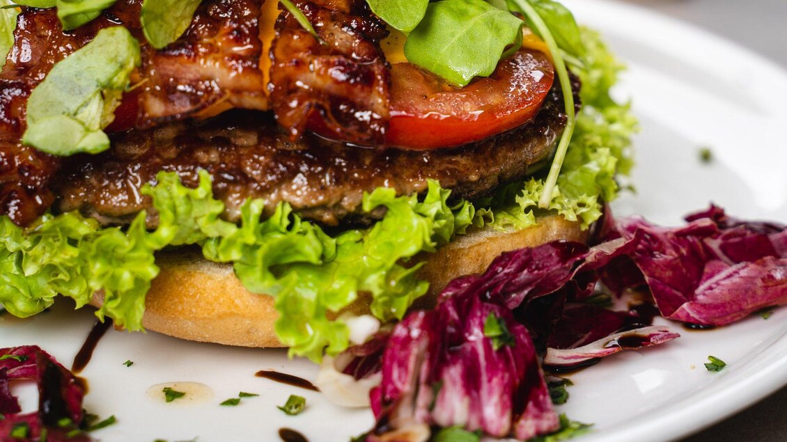 Ein saftiger Burger mit Bacon, Salat und Tomaten. Dazu frischer Radicchio auf einem weißen Teller. | © Arvideo
