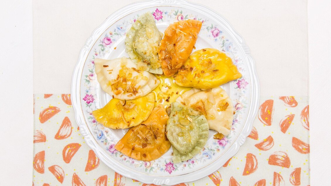 Ein Teller mit verschiedenen gefüllten Teigtaschen in verschiedenen Farben. Die Teigtaschen sind mit einer goldbraunen Füllung und leicht angeröstetem Zwiebelbelag dekoriert. | © Pierogi