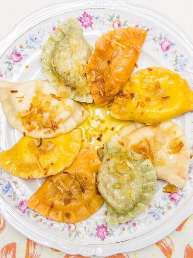 A plate with various filled dumplings in different colors. The dumplings are decorated with a golden-brown filling and a lightly roasted onion topping. | © Pierogi