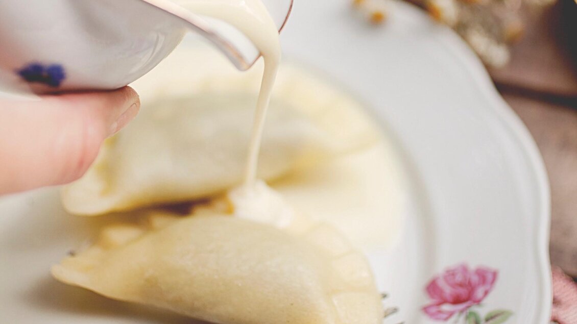 Eine Hand gießt eine cremige Sauce aus einer Kanne auf einen Teller mit einem Gericht. Im Hintergrund sind Blumen zu sehen. | © Pierogi
