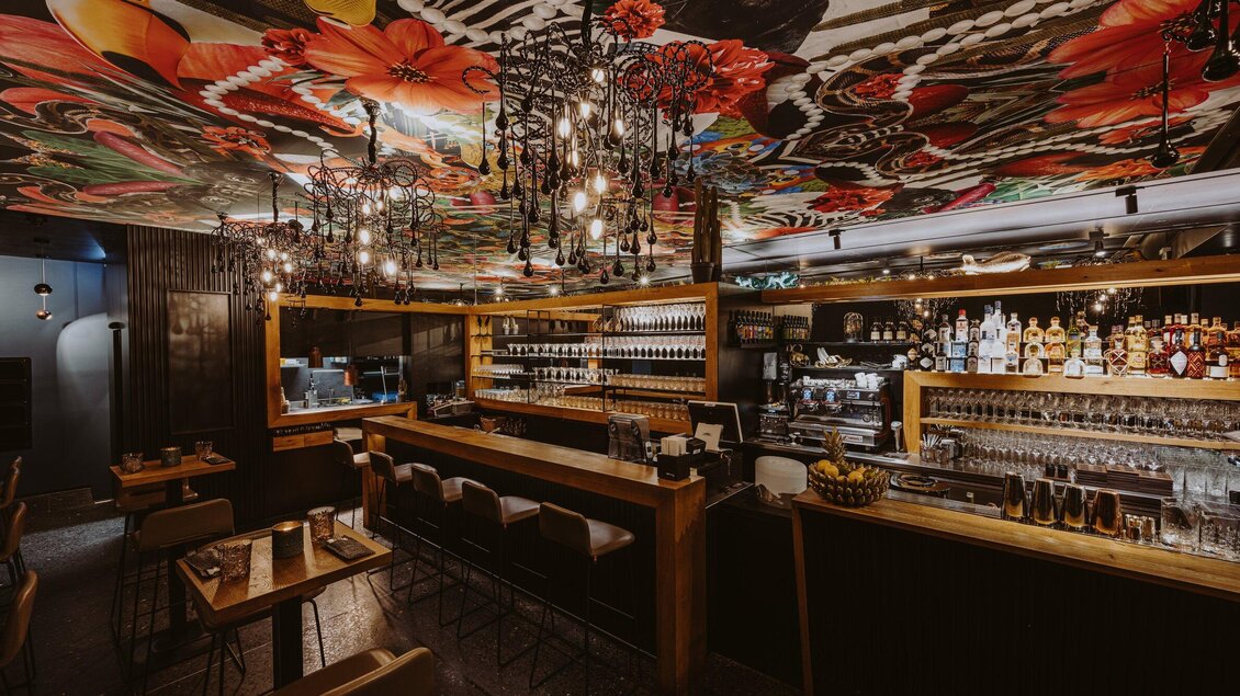 Ein stilvolles Restaurant mit einer langen Bar und eleganter Beleuchtung. Die Decke ist mit bunten Blumenmustern dekoriert und schafft eine einladende Atmosphäre. | © Aiola - Joel Kernasenko