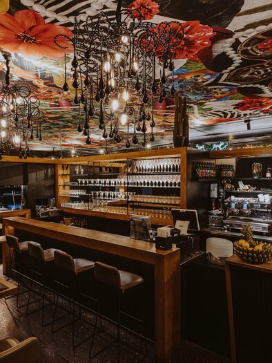 A stylish restaurant with a long bar and elegant lighting. The ceiling is adorned with colorful floral patterns, creating an inviting atmosphere. | © Aiola - Joel Kernasenko