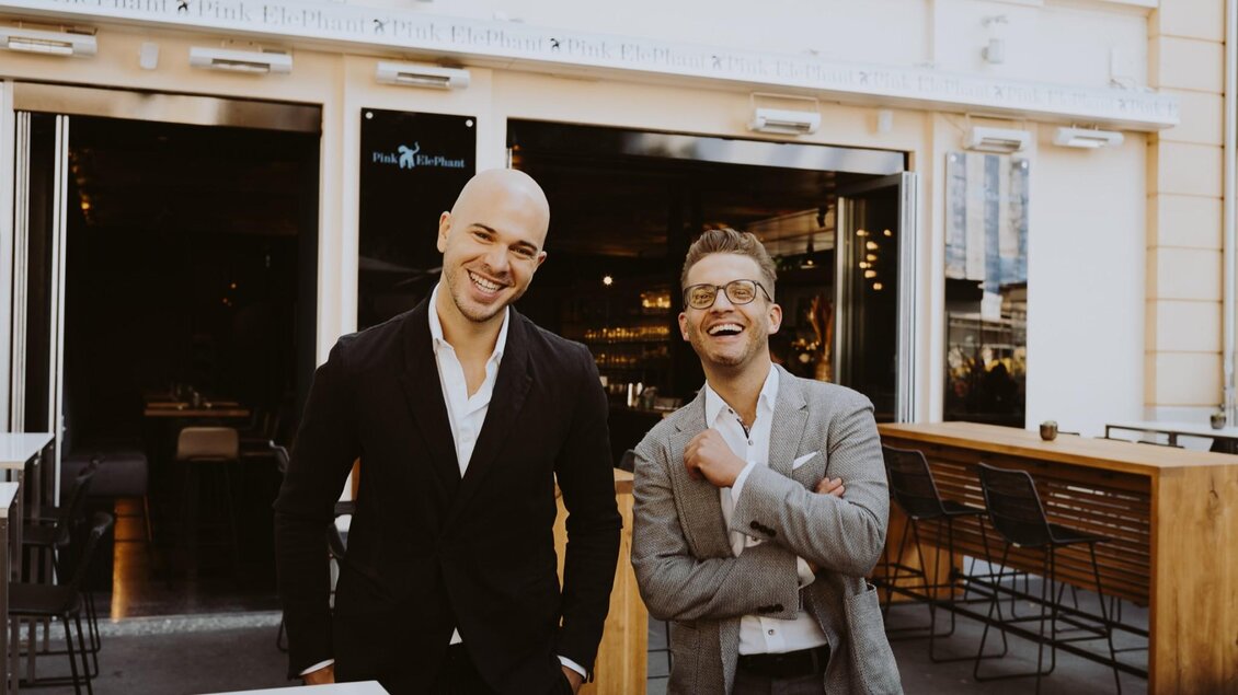 Zwei Männer lächeln freundlich vor einem Restaurant. Sie tragen elegante Anzüge und stehen vor einer hellen, modernen Fassade. | © Aiola - Joel Kernasenko