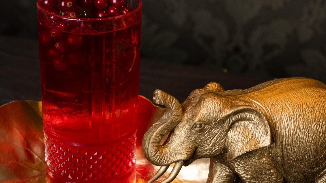 Ein Glas mit rotem Getränk steht auf einem goldenen Tablett, neben einem goldenen Elefanten. Die Atmosphäre ist elegant und einladend. | © Aiola - Jauk Katharina