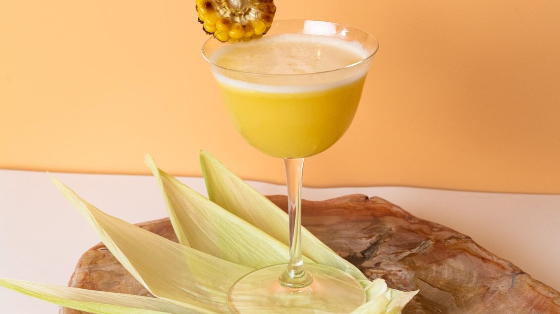 Ein erfrischender Cocktail in einem eleganten Glas, dekoriert mit Ananas. Er steht auf einem Holzbrett, umgeben von Ananasblättern. | © Aiola - Jauk Katharina