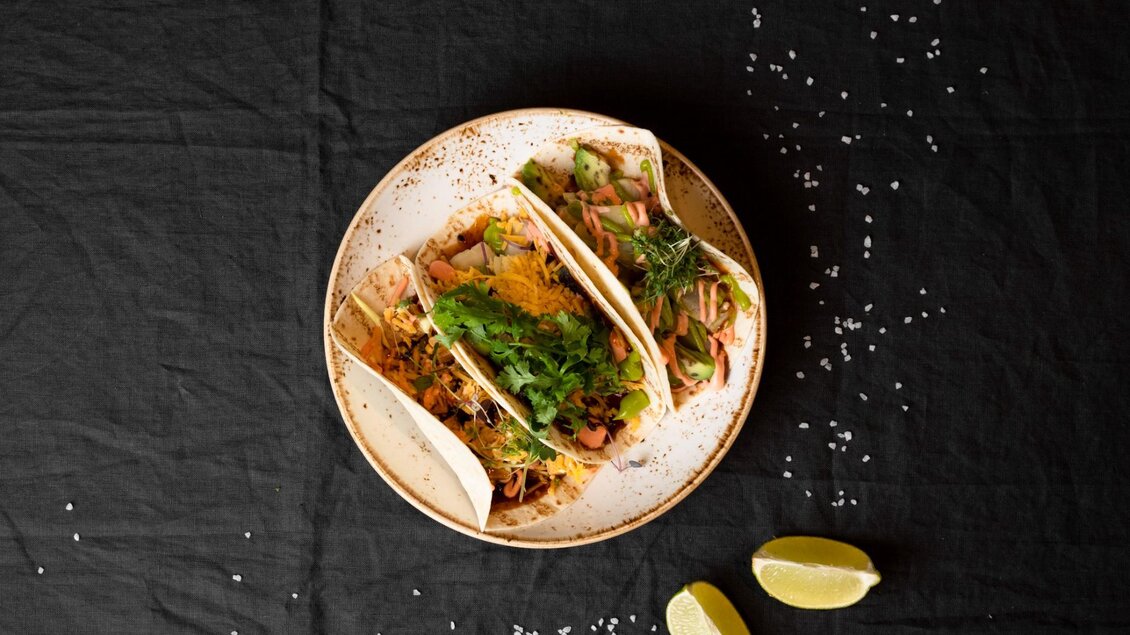 Ein Teller mit drei leckeren Tacos, gefüllt mit frischen Zutaten und Kräutern. Neben dem Teller liegen zwei Limetten. | © Aiola - Jauk Katharina