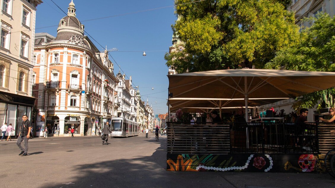 Eine belebte Straßenszene mit schönen historischen Gebäuden und einer Terrasse eines Cafés. Die Sonne scheint und Menschen gehen spazieren. | © Aiola - Jauk Katharina