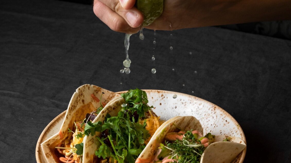 Ein Teller mit drei gefüllten Tacos, garniert mit frischen Kräutern. Eine Hand träufelt Limettensaft über die Tacos. | © Aiola - Jauk Katharina