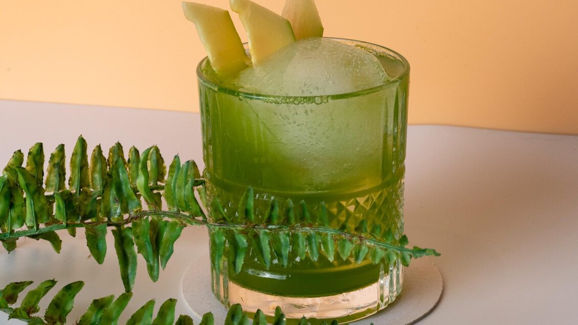 Ein erfrischendes grünes Getränk in einem eleganten Glas mit Eis und Früchten. Im Vordergrund sind grüne Blätter zu sehen. | © Aiola - Jauk Katharina