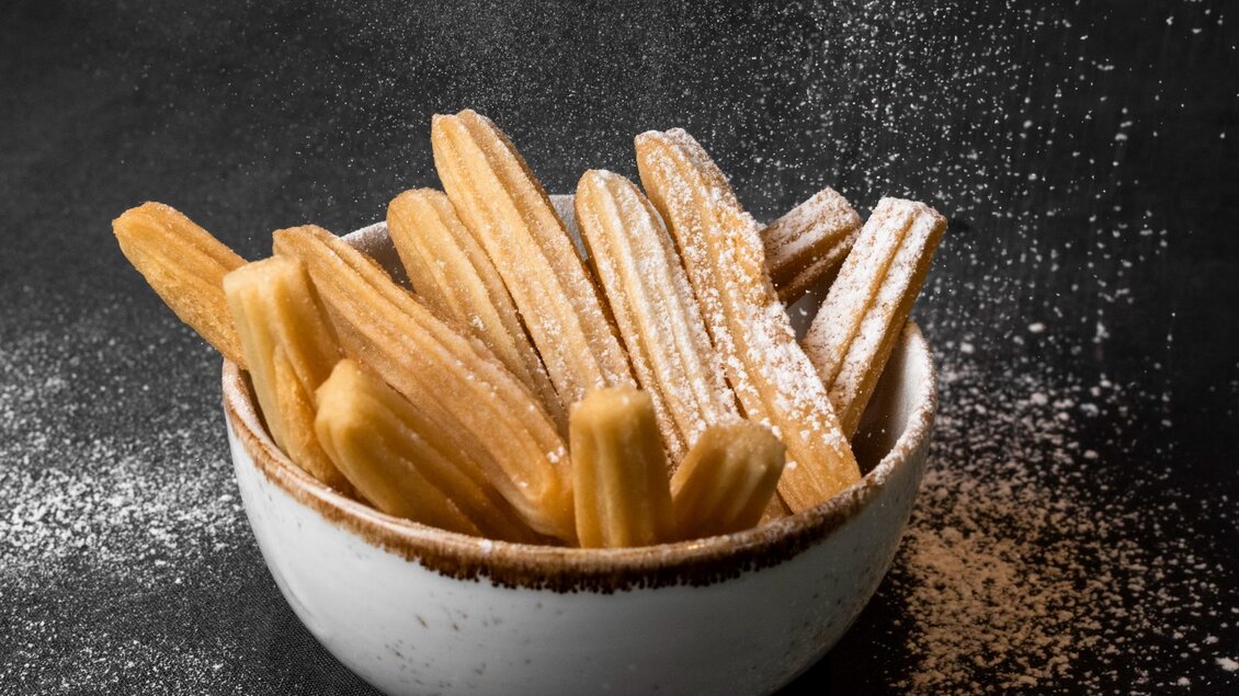 Ein Teller mit frischen Churros, die mit Puderzucker bestäubt sind. Die Churros haben eine goldene Farbe und sind appetitlich angerichtet. | © Aiola - Jauk Katharina
