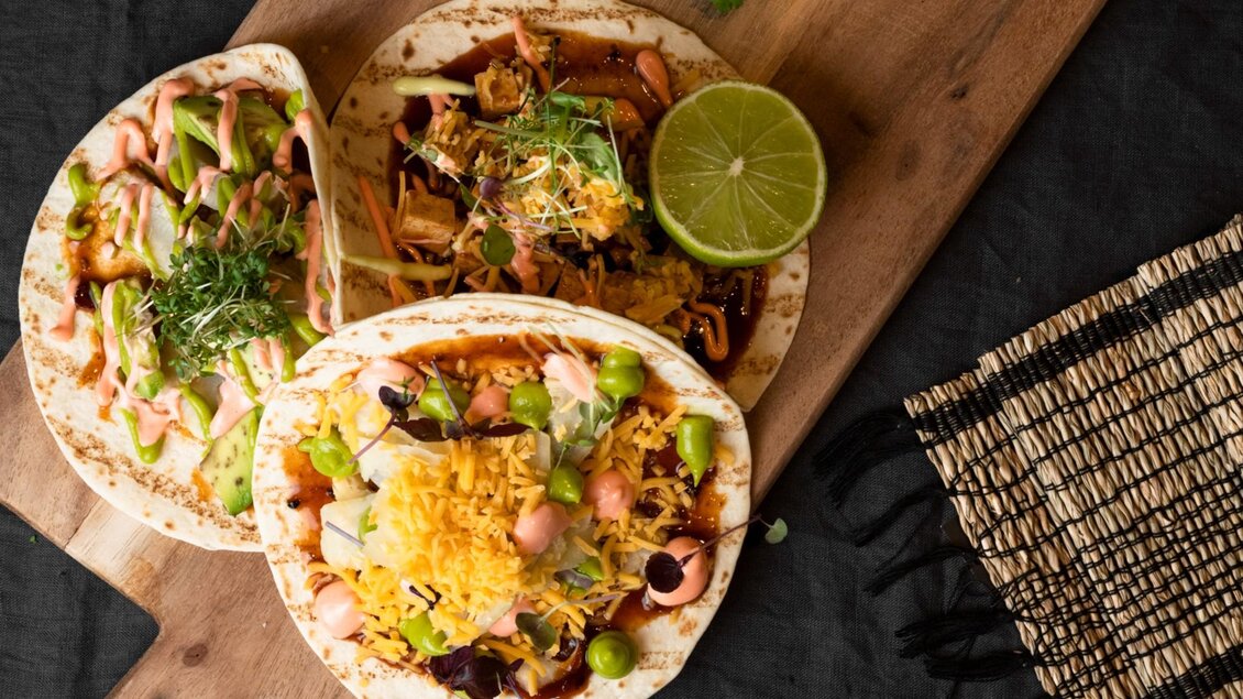 Drei leckere Tacos auf einem Holzbrett, garniert mit frischem Gemüse und Käse. Dazu liegt eine Limette für zusätzlichen Geschmack. | © Aiola - Jauk Katharina