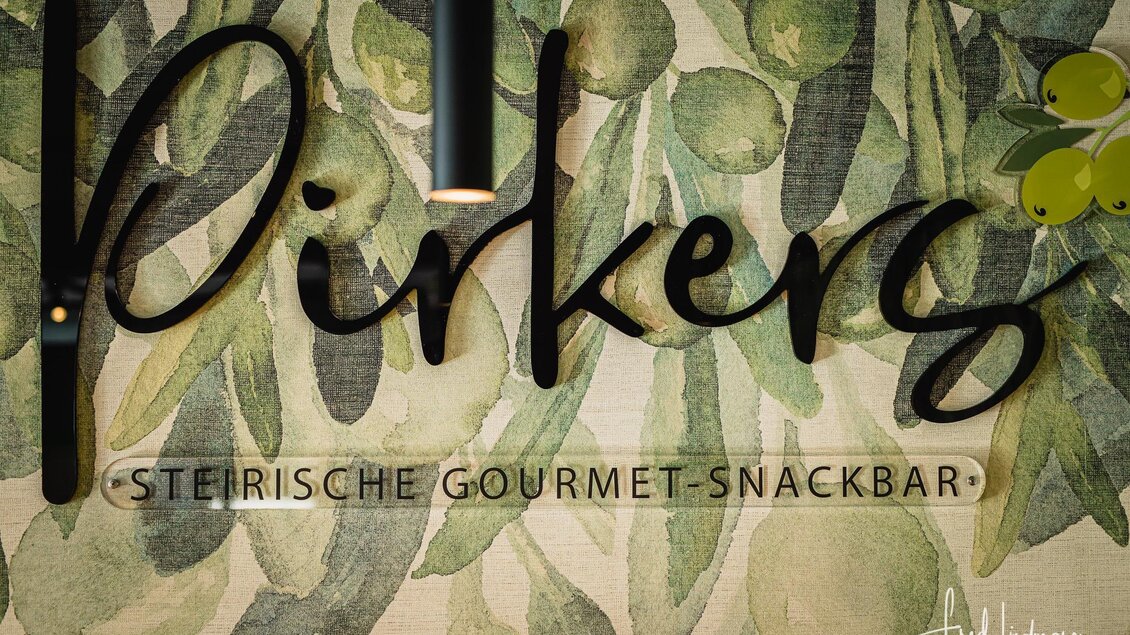 Ein dekoratives Schild mit dem Namen "Pirker's" und dem Untertitel "Steirische Gourmet-Snackbar". Der Hintergrund zeigt ein florales Muster mit Oliven und Blättern. | © Pirker GmbH Mariazell