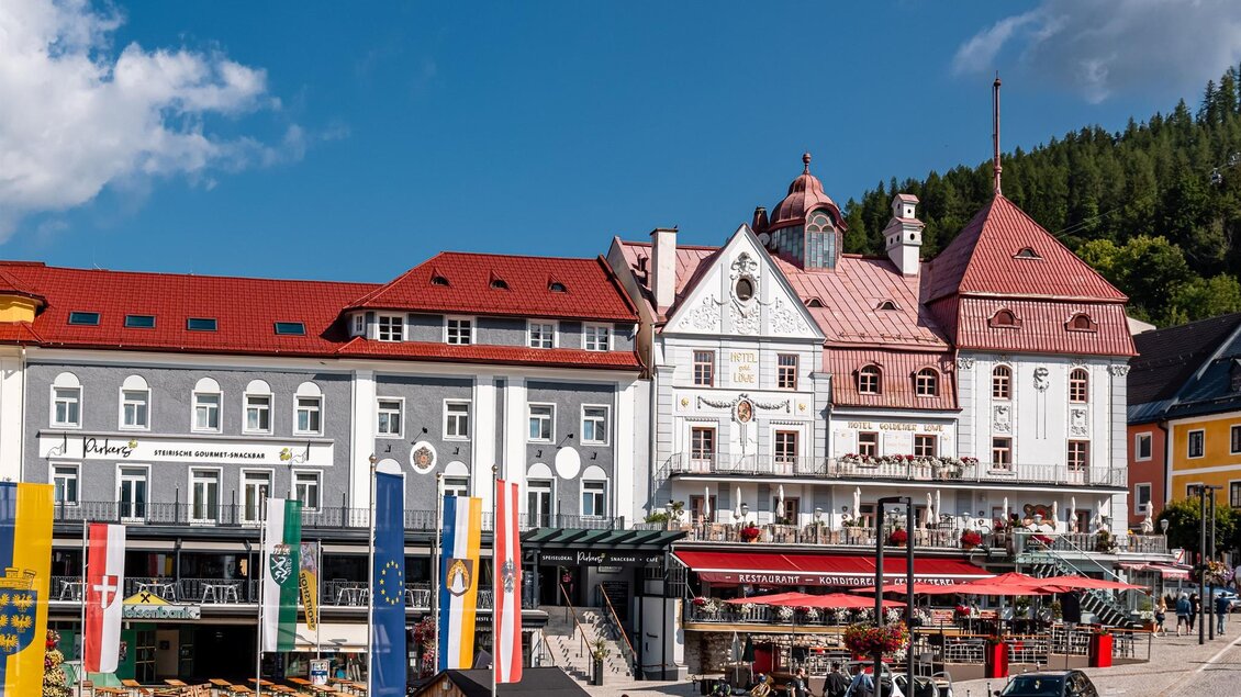 Eine charmante Stadtansicht mit historischen Gebäuden und einem Restaurant im Freien. Im Hintergrund sind grüne Hügel und ein blauer Himmel zu sehen.