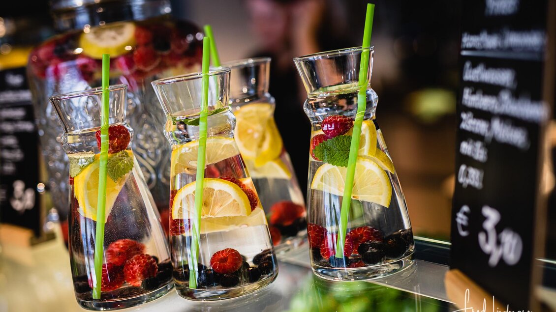 Eine Auswahl an erfrischenden Getränken in Glasbehältern, gefüllt mit Limonen, Zitronen und Beeren. Die Gläser stehen auf einem Tisch neben einer Menütafel. | © www.mariazell.blog / Fred Lindmoser