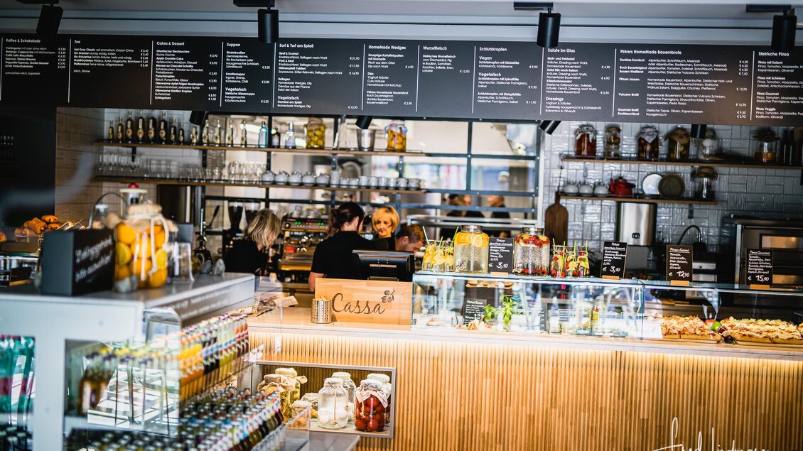 Ein modernes Café mit einem hellen Thekenbereich. Im Hintergrund sind Mitarbeiter und eine Auswahl an Getränken und Snacks zu sehen. | © www.mariazell.blog / Fred Lindmoser