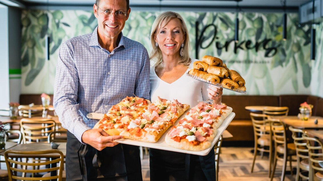 Zwei Personen stehen in einem Restaurant und halten eine große Platte mit Pizza und Gebäck. Im Hintergrund sind Tische und eine grüne Wandgestaltung zu sehen. | © Pirker GmbH Mariazell