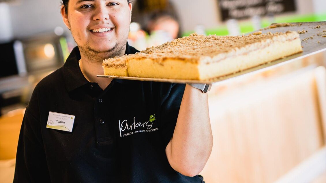 Ein freundlicher Mitarbeiter hält ein großes Stück Kuchen hoch. Im Hintergrund sind die Menüliste und eine moderne Café-Atmosphäre zu sehen. | © Pirker GmbH Mariazell