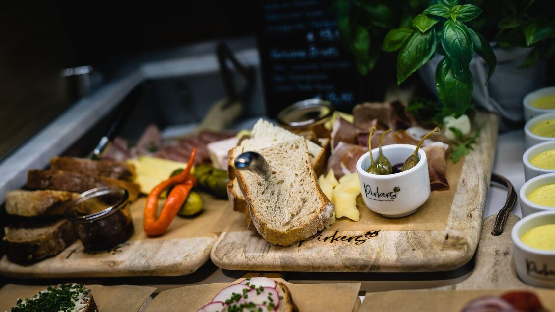 Ein reichhaltiges Grillbuffet mit verschiedenen Wurst- und Käsesorten, frischem Brot und buntem Gemüse. Auf einem Holzbrett angerichtet, daneben Tassen mit Dip. | © Pirker GmbH Mariazell