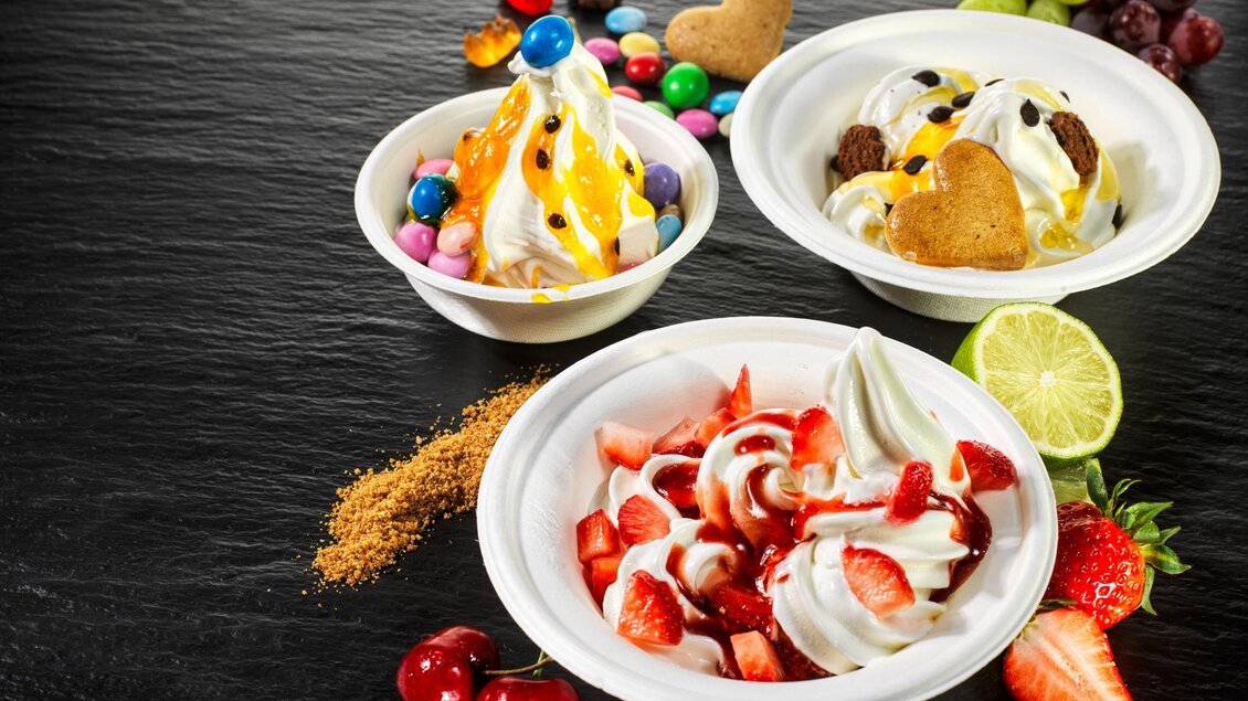 Drei Becher mit köstlichem Frozen Yogurt, dekoriert mit bunten Süßigkeiten und frischen Früchten. Dazu liegen einige Früchte und Zucker auf einer dunklen Unterlage. | © Pirker GmbH Mariazell