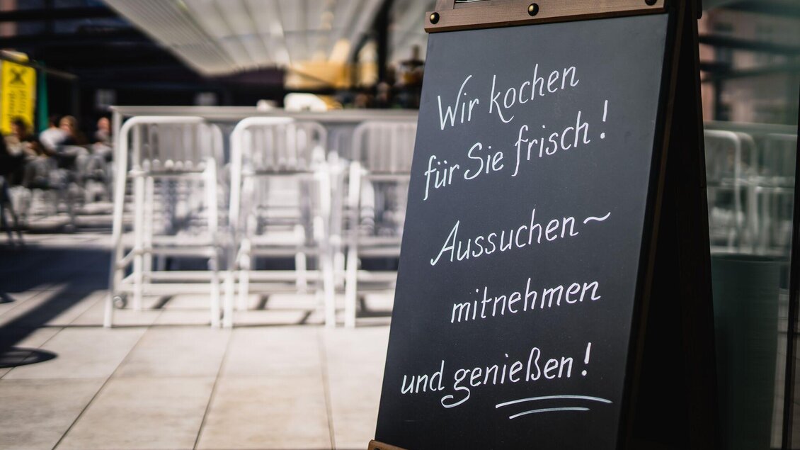 Ein schwarzes Schild mit weißer Schrift, das Werbung für frisches Essen macht. Der Text lädt ein, die Speisen auszuwählen, mitzunehmen und zu genießen. | © Pirker GmbH Mariazell