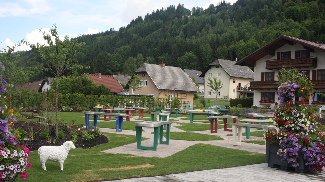 Eine schöne Gartenlandschaft mit bunten Tischen und Stühlen. Im Hintergrund sind traditionelle Häuser und grüne Hügel zu sehen. | © TVB Haus-Aich-Gössenberg