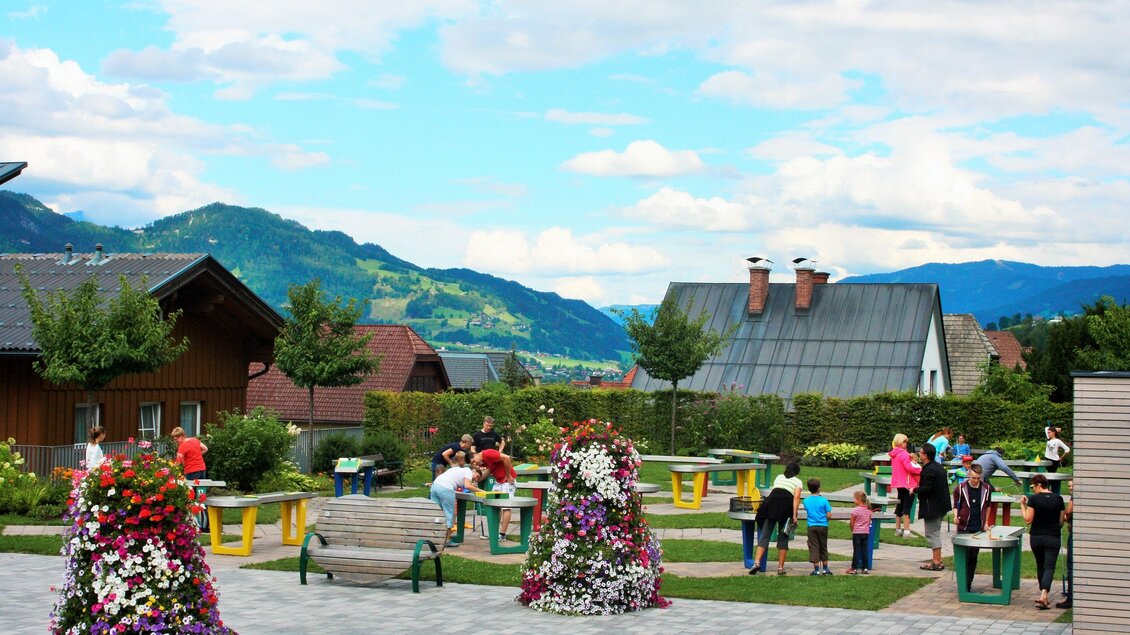 Ein schöner Park mit bunten Blumen und mehreren Menschen, die sich entspannen und spielen. Im Hintergrund sind Berge und Häuser zu sehen. | © TVB Haus-Aich-Gössenberg