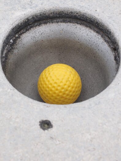A yellow golf ball lies in a concrete hole. The background is gray and plain. | © Puttererseehof