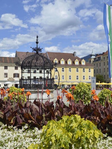 Ein schöner Platz mit bunten Blumen und einer dekorativen Brunnenskulptur. Im Hintergrund sind historische Gebäude und Fahnen zu sehen. | © M. Maili