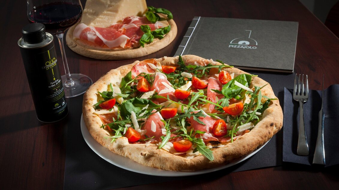 Eine köstliche Pizza mit Rucola, Schinken, Tomaten und Parmesan. Dazu ein Glas Wein und ein Menübuch auf dem Tisch. | © Pizzaiolo