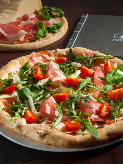 A delicious pizza with arugula, ham, tomatoes, and Parmesan. Accompanied by a glass of wine and a menu book on the table. | © Pizzaiolo