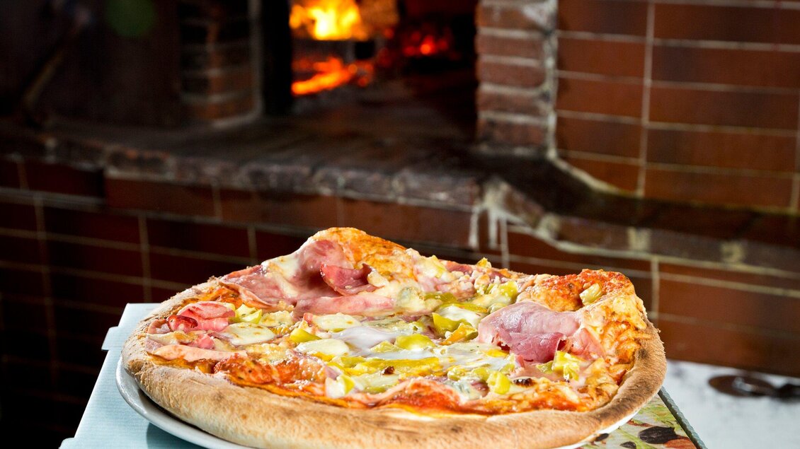 Eine frisch gebackene Pizza mit Schinken, Käse und Gemüse. Im Hintergrund sieht man einen Holzofen. | © TV Region Graz - René Vidalli