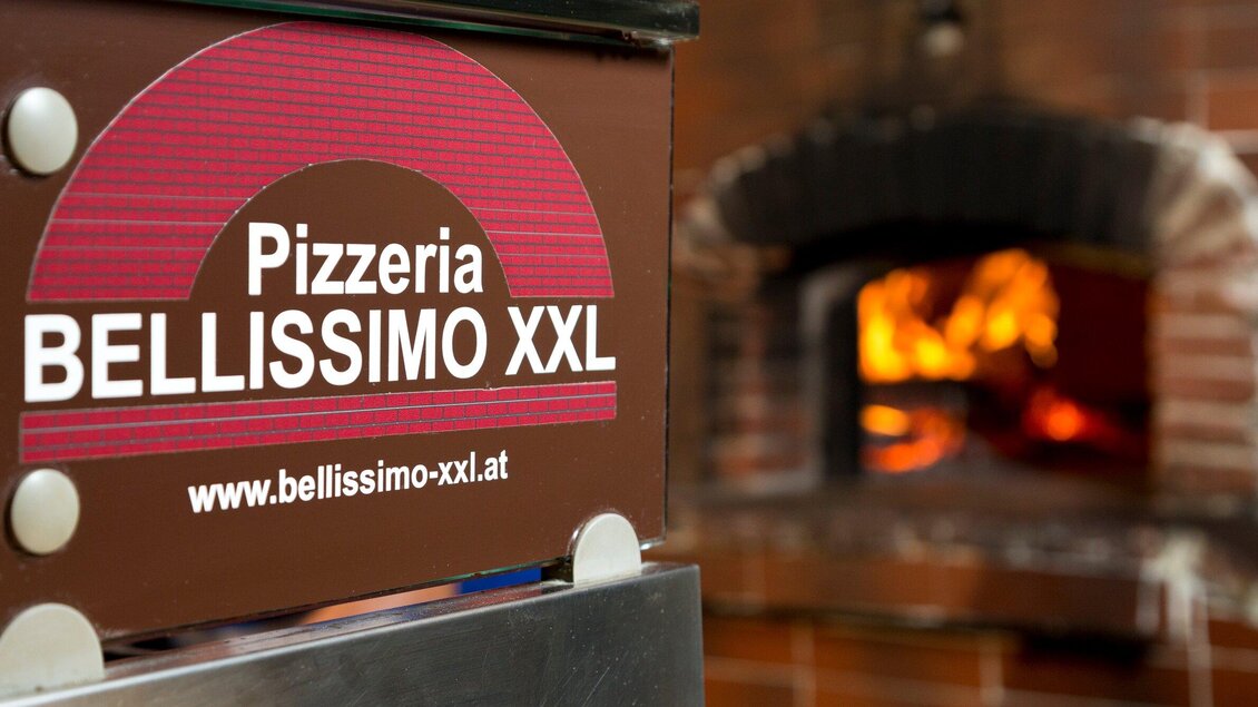 Ein Schild der Pizzeria Bellissimo XXL steht neben einem offenen Holzofen. Der Ofen ist mit flammendem Feuer gefüllt, was eine gemütliche Atmosphäre erzeugt. | © TV Region Graz - René Vidalli