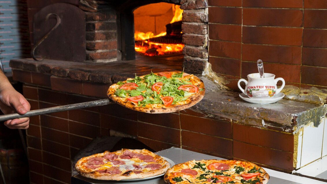 Eine Pizzaschaufel hebt eine frisch gebackene Pizza mit Salat und Tomaten. Im Hintergrund ist ein traditioneller Holzofen zu sehen. | © TV Region Graz - René Vidalli