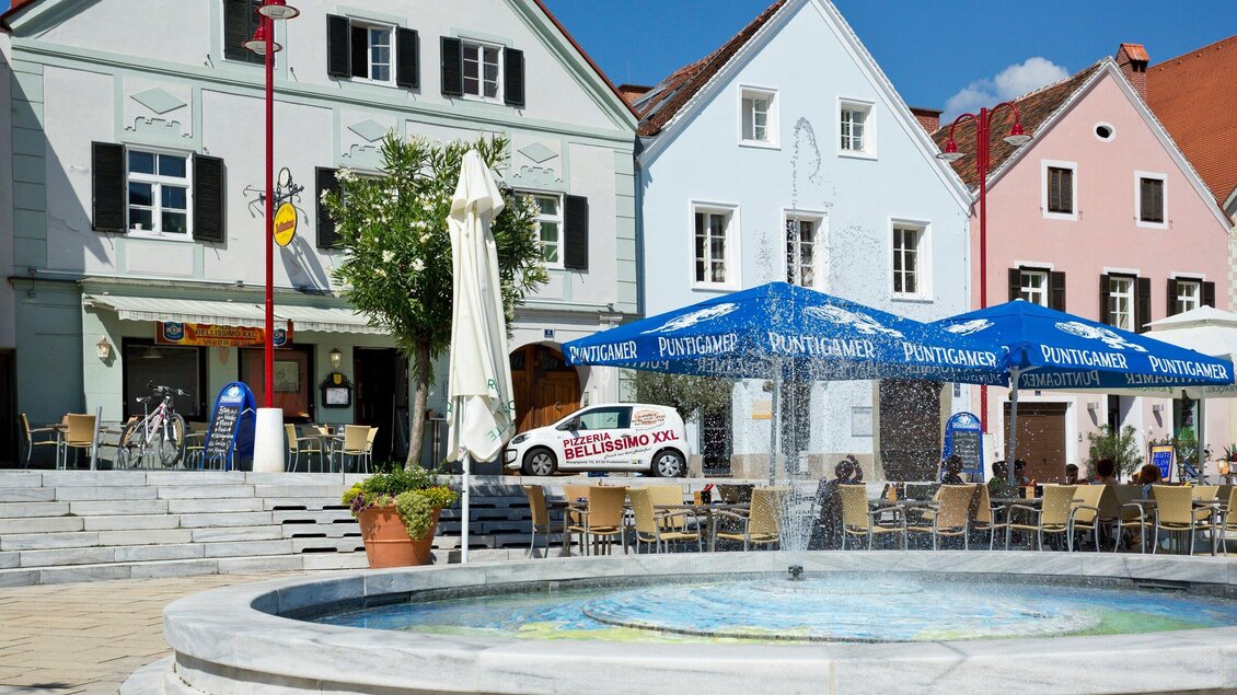 Ein schöner Platz mit einem Brunnen und bunten Gebäuden. Es gibt Stühle und Tische unter einem Sonnenschirm. | © TV Region Graz - René Vidalli