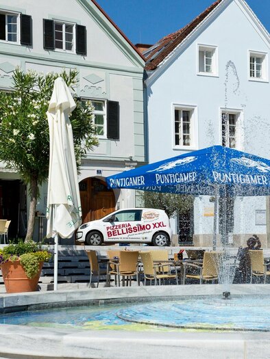 Ein schöner Platz mit einem Brunnen und bunten Gebäuden. Es gibt Stühle und Tische unter einem Sonnenschirm. | © TV Region Graz - René Vidalli