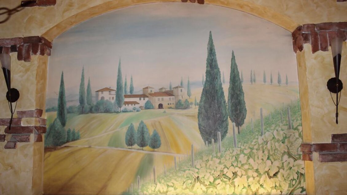 Ein malerisches Wandgemälde mit einer Landschaft, die sanfte Hügel, Zypressen und ein typisches italienisches Dorfbild zeigt. Die Farben sind sanft und harmonisch, was eine ruhige Atmosphäre schafft. | © Pizzeria Fratelli
