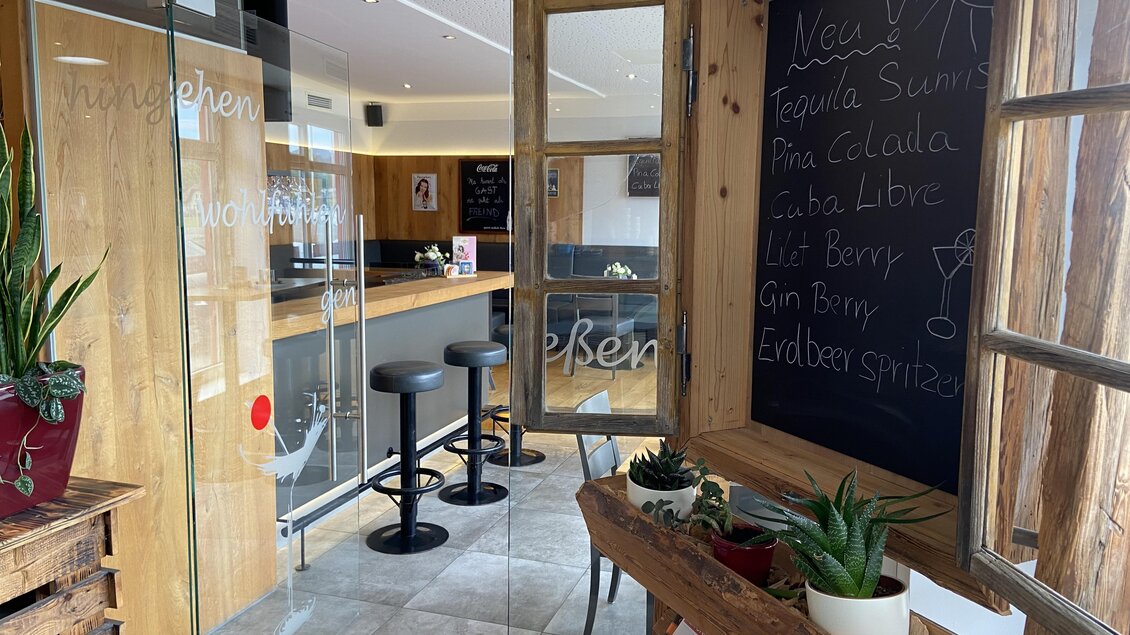 Ein einladender Innenraum mit einem Holzregal und Pflanzen. Im Hintergrund ist eine Bar mit Barhockern und einer Tafel mit Getränkespecials zu sehen. | © Mamarosa