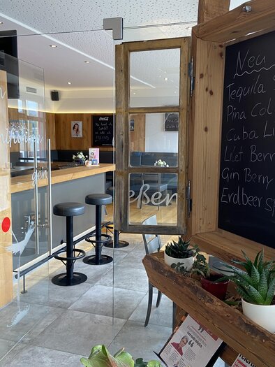An inviting interior with a wooden shelf and plants. In the background, there is a bar with bar stools and a chalkboard displaying drink specials. | © Mamarosa