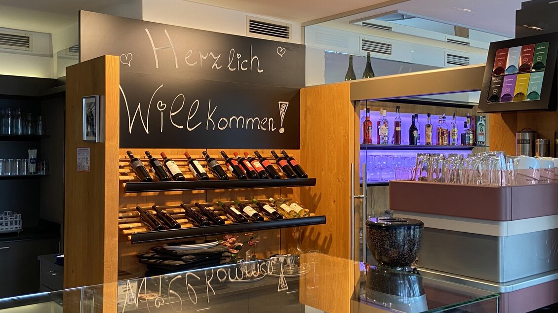 Ein einladendes Café mit einem Holzregal voller Weinflaschen. An der Wand steht "Herzlich Willkommen!" in geschwungener Schrift. | © Mamarosa