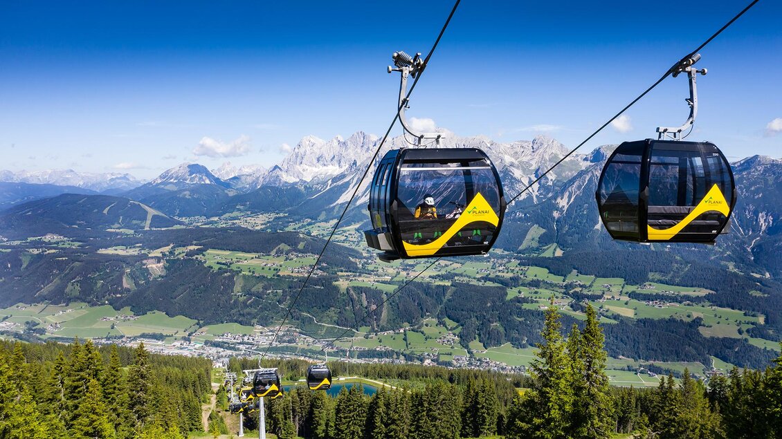Eine Seilbahn fährt über eine beeindruckende Berglandschaft. Die Aussicht zeigt grüne Täler und hohe Berge unter einem klaren blauen Himmel. | © Josh Absenger