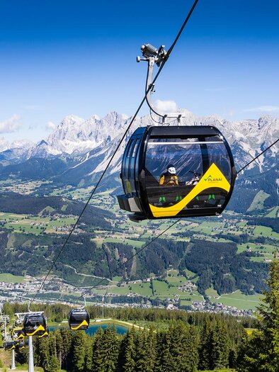 A cable car travels over an impressive mountain landscape. The view shows green valleys and high mountains under a clear blue sky. | © Josh Absenger