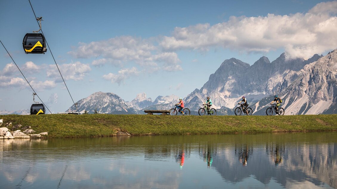 Eine malerische Landschaft mit Radfahrern am Ufer eines Sees und beeindruckenden Bergen im Hintergrund. Eine Seilbahn schwebt über die Szenerie und fügt sich harmonisch in die Natur ein. | © Roland Haschka