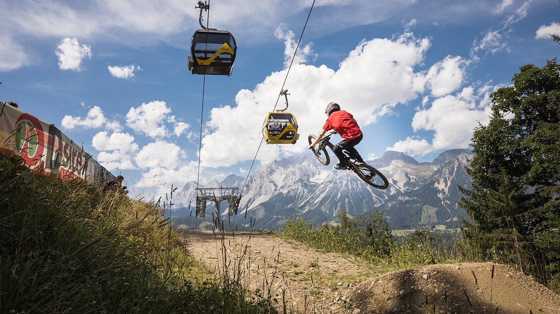 Ein Mountainbiker springt durch die Luft auf einem Weg in den Bergen. Im Hintergrund sind Seilbahnen und eine malerische Berglandschaft sichtbar. | © Roland Haschka